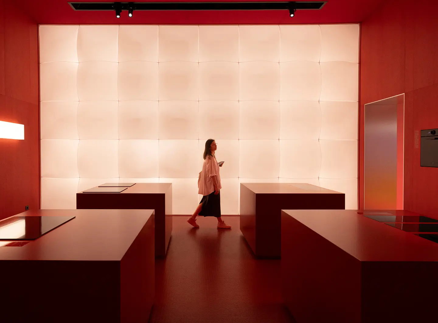 Perspective view of the stand with a girl in the centre and four counters on the sides. In the background, a panel of lights illuminating the room, the colours of which tend towards red.