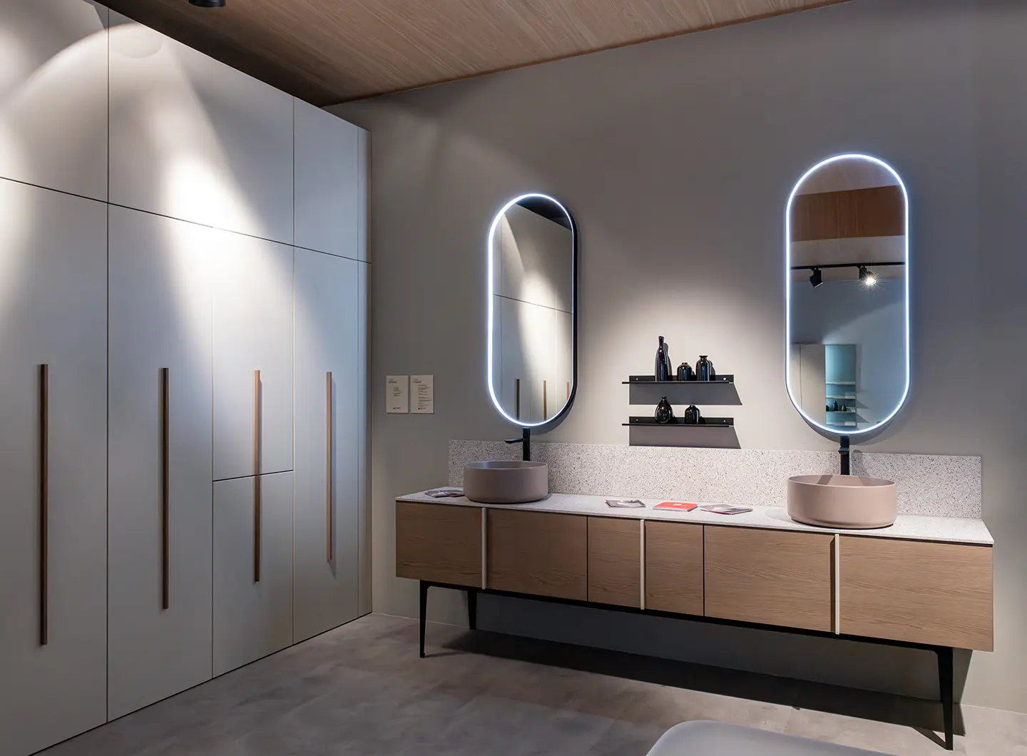 Mobile bagno in legno chiaro con due lavabi rotondi in ceramica e due specchi ovali, con mobili bianchi a tutta altezza sul lato. 