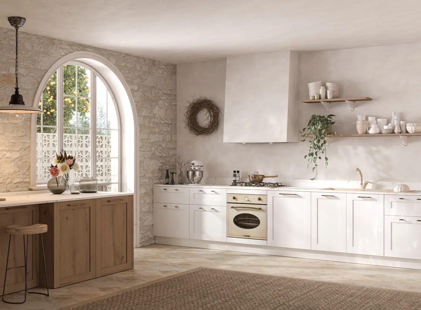 Ampia cucina in stile rustico situata in una stanza con pareti in pietra a vista e una grande finestra ad arco. La cucina presenta basi bianche con maniglie a conchiglia color ottone, con un forno 60 cm e un piano 70 cm dall’estetica tradizionale color avena e una cappa integrata aspirante.