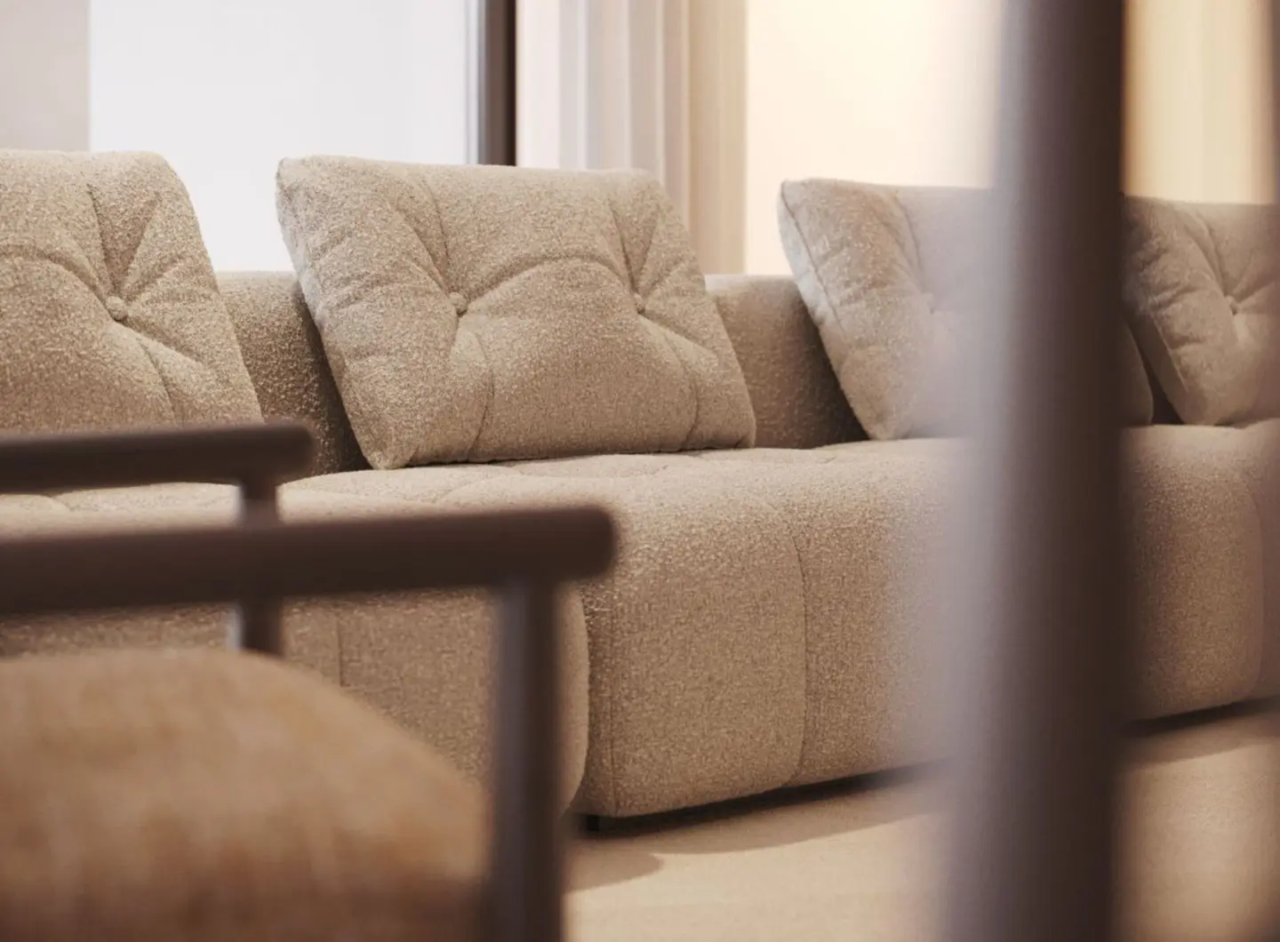Il divano Antónia è mostrato in primo piano come un modulo beige in bouclé con cuscini profondamente trapuntati e una texture morbida, accompagnato da una poltrona coordinata con braccioli in legno in primo piano, inserito in un ambiente dai toni neutri che esalta la ricchezza tattile e il design minimalista del pezzo.
