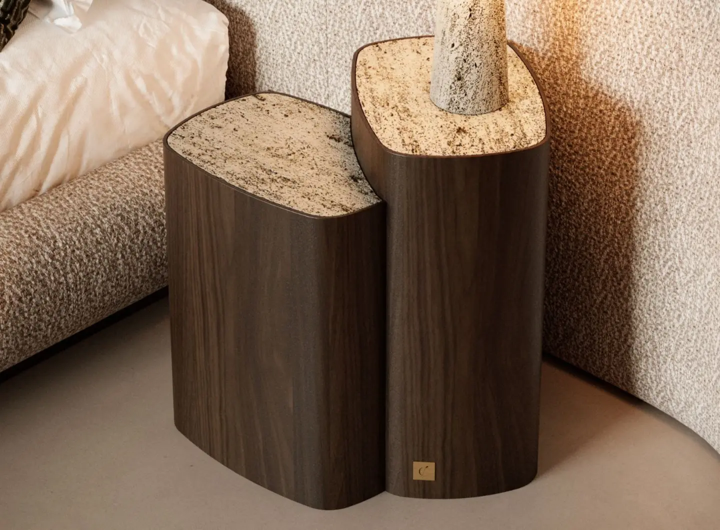 Il Tavolino di servizio Dolomites è mostrato come una coppia di tavolini in legno con una base curva e organica e piani in pietra lucida nei toni del beige e del marrone, posizionati accanto a un letto, con uno dei tavolini che sostiene un oggetto cilindrico in pietra e una piccola targhetta in ottone visibile vicino alla base.
