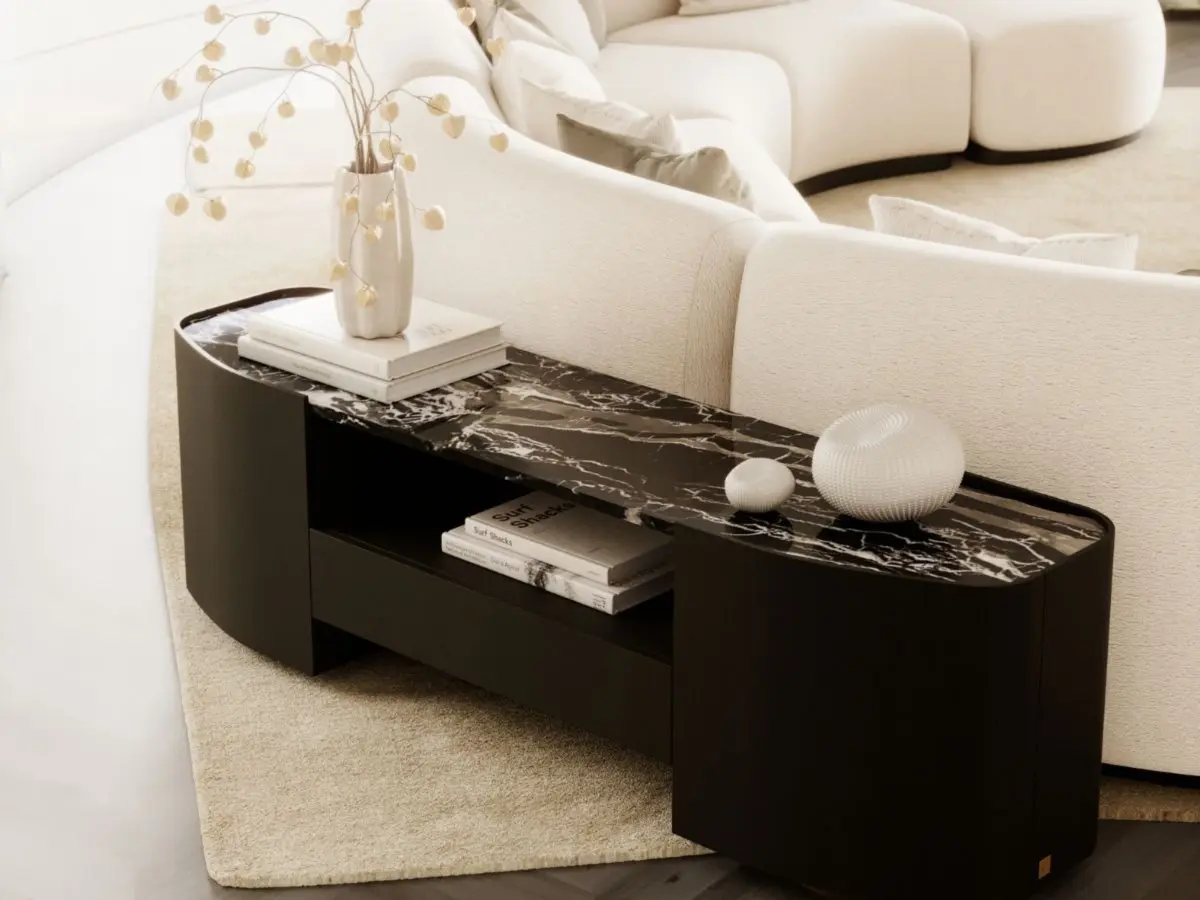 La consolle bassa Stone, con piano in pietra nera lucida e base arrotondata in legno scuro, è collocata dietro un divano curvo color crema, impreziosito da un vaso bianco, elementi decorativi scultorei e libri impilati, in un salotto dai toni tenui e neutri.